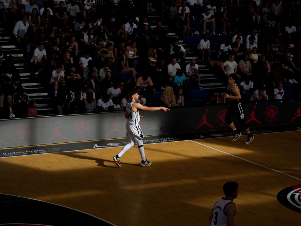 A basketball player standing in the light in a shadowed basketball gym during the 2023 Hoopsfix All-Star Classic. A picture taken by the Sports Media Agency Corzek.